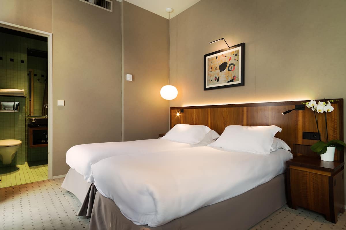 A hotel room with two beds, a wooden headboard, a bedside table with a plant, a framed picture on the wall, and a bathroom visible through an open door.
