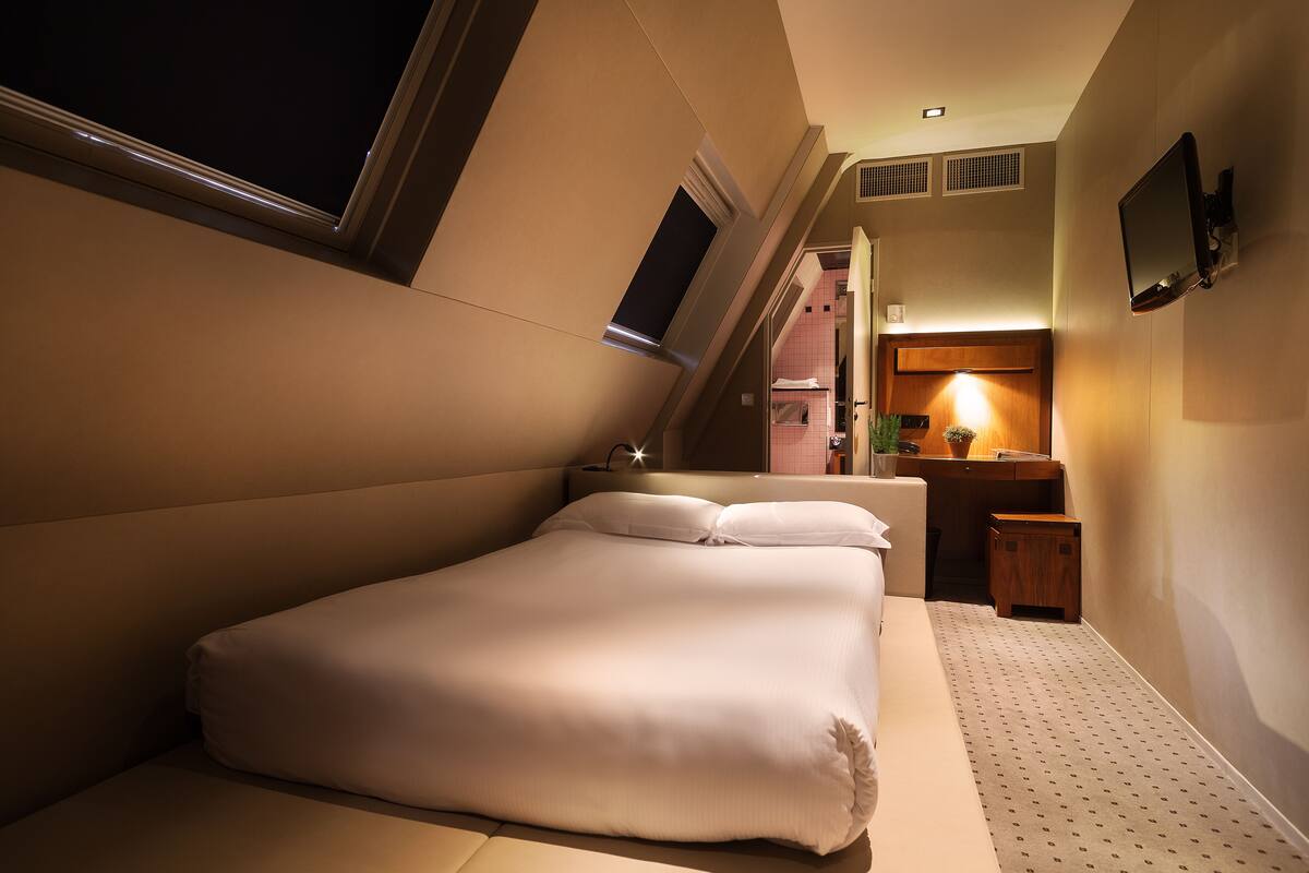 A hotel room with a large bed, a small desk, a nightstand, and a wall-mounted TV.