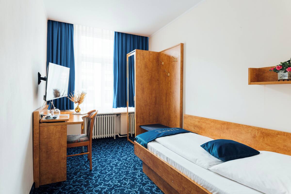 A room with a bed, a desk, a chair, a radiator, and a window with curtains.