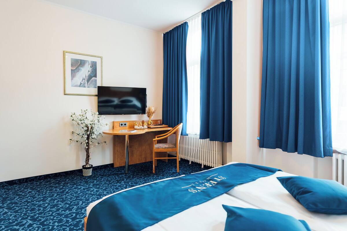 A hotel room with a bed, a desk, a chair, a TV, and a potted plant.