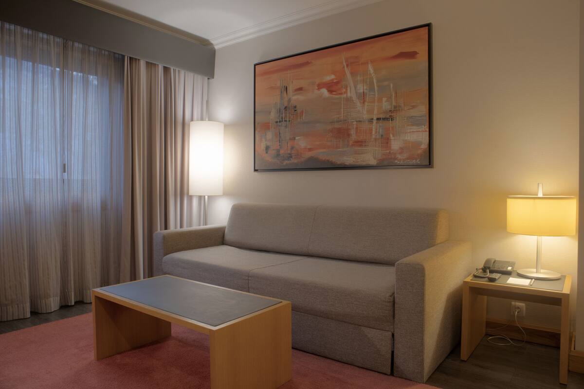 A hotel room with a grey sofa, a wooden coffee table, a lamp on a bedside table, and a large abstract painting on the wall.