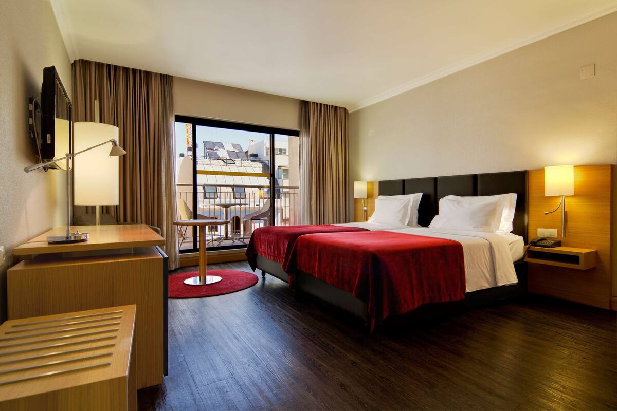 A hotel room with a bed, bedside tables, a desk, a chair, and a large window with curtains.