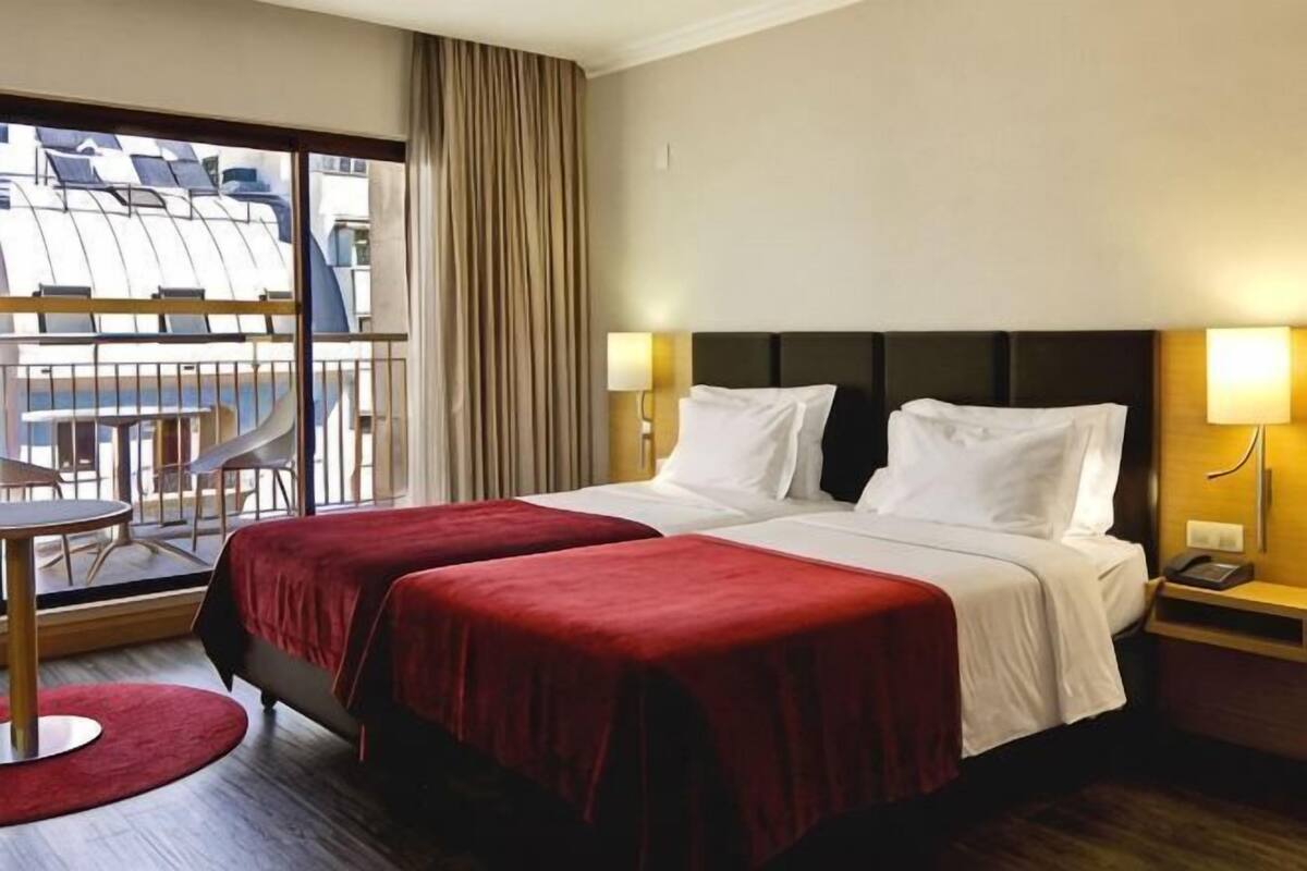 A hotel room with two beds, a balcony with a view, a round table, and a chair.