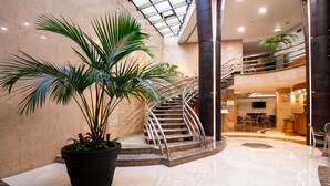 Eine großzügige Lobby mit einer hohen Pflanze in einem Topf, einer geschwungenen Treppe und einer Sitzecke mit Tischen und Stühlen.