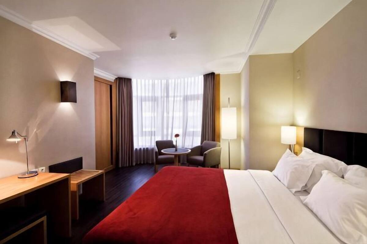 A hotel room with a bed, a desk, a chair, a small table, and a lamp.