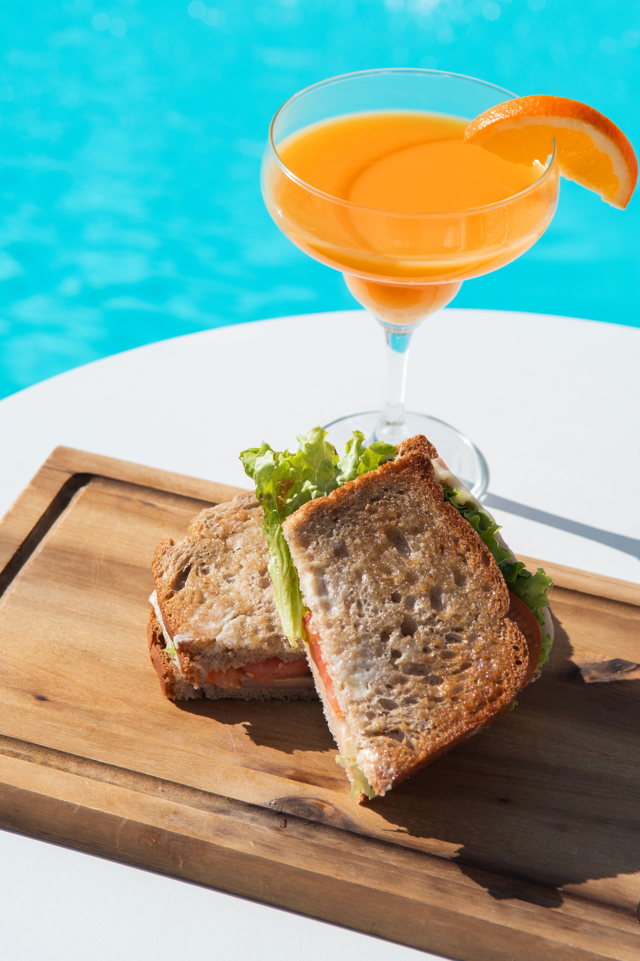 Un sándwich sobre una tabla de madera, con un cóctel al fondo.