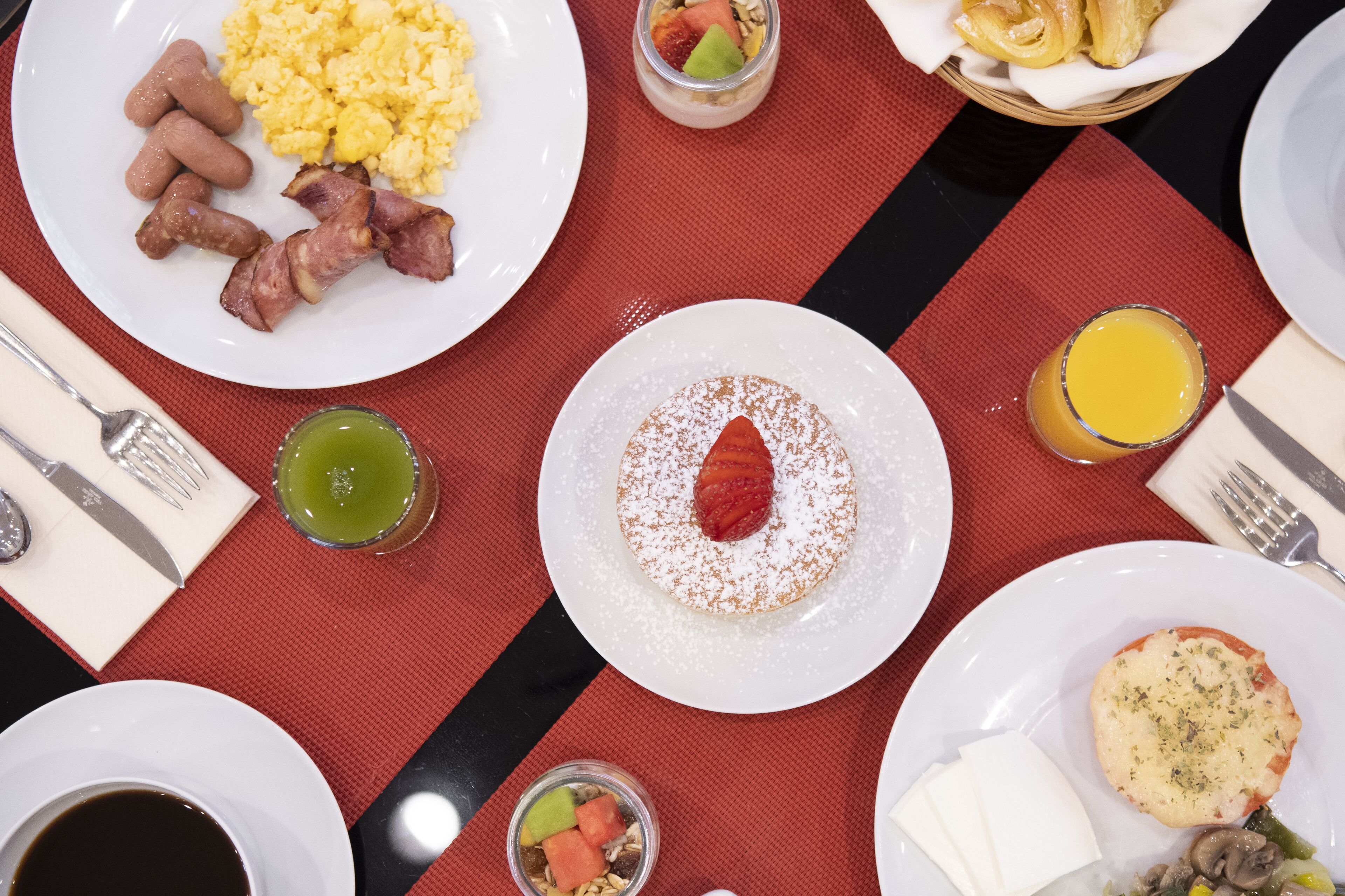 A breakfast setting with plates of scrambled eggs, sausages, bacon, a fruit salad, a strawberry dessert, a glass of orange juice, and a cup of coffee.
