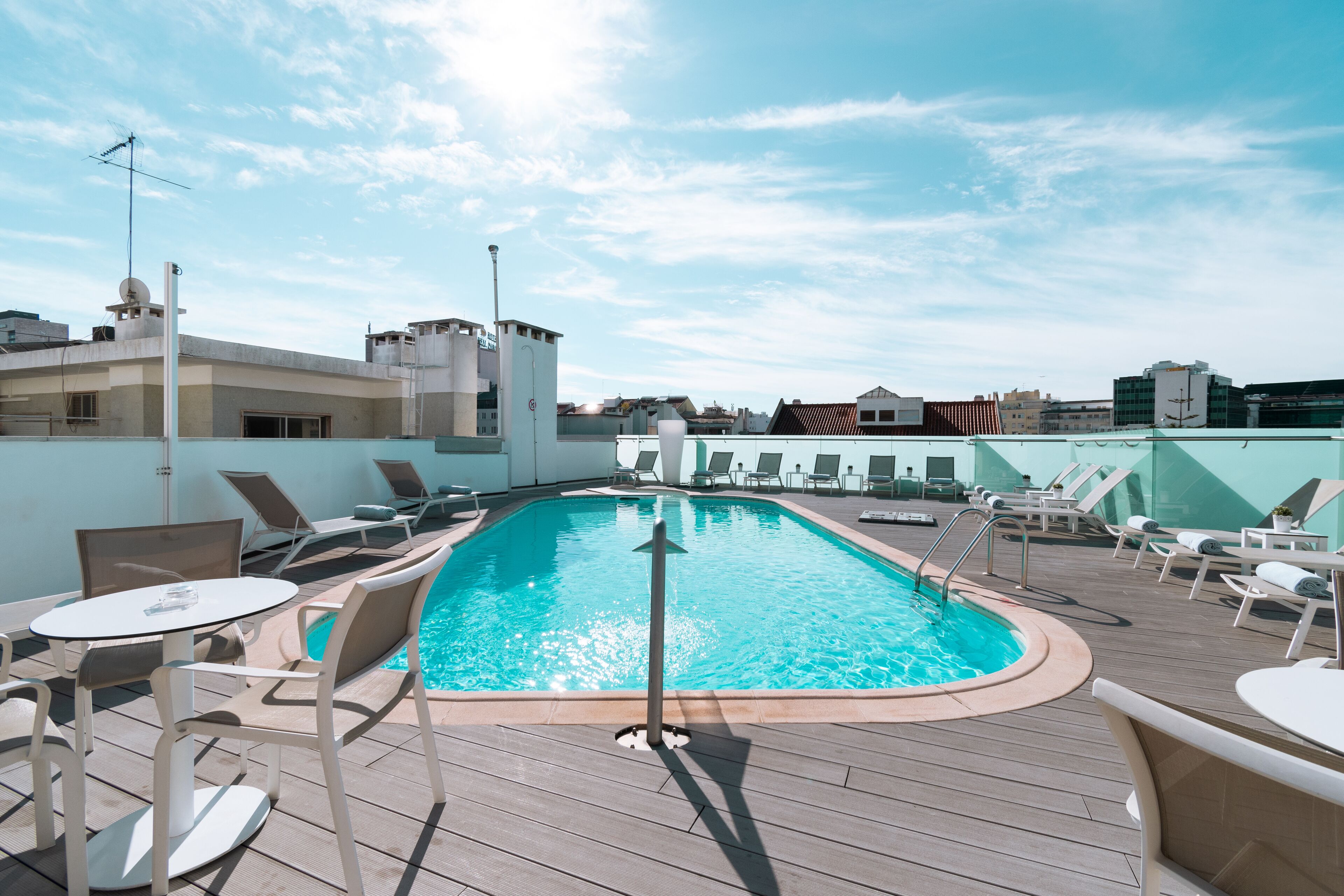 Un área de piscina en la azotea con sillas de descanso y una mesa, rodeada de edificios.