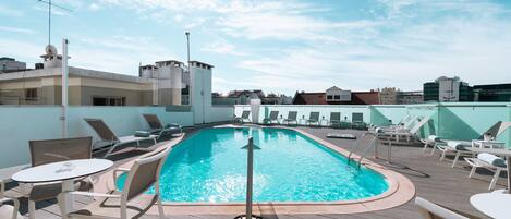 Un área de piscina en la azotea con sillas de descanso y una mesa, rodeada de edificios.