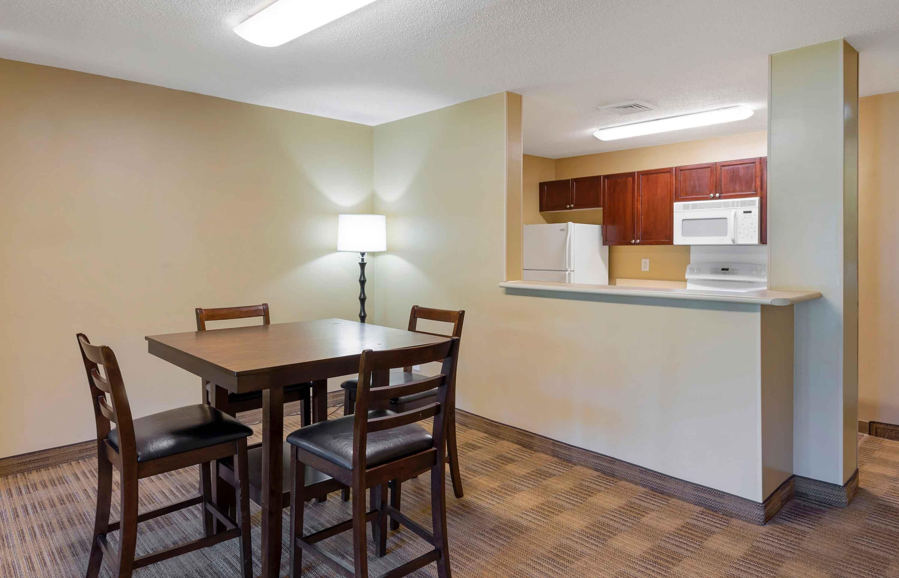 A compact hotel room with a dining table, four chairs, a small kitchenette, and a single floor lamp.