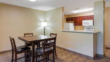 A compact hotel room with a dining table, four chairs, a small kitchenette, and a single floor lamp.