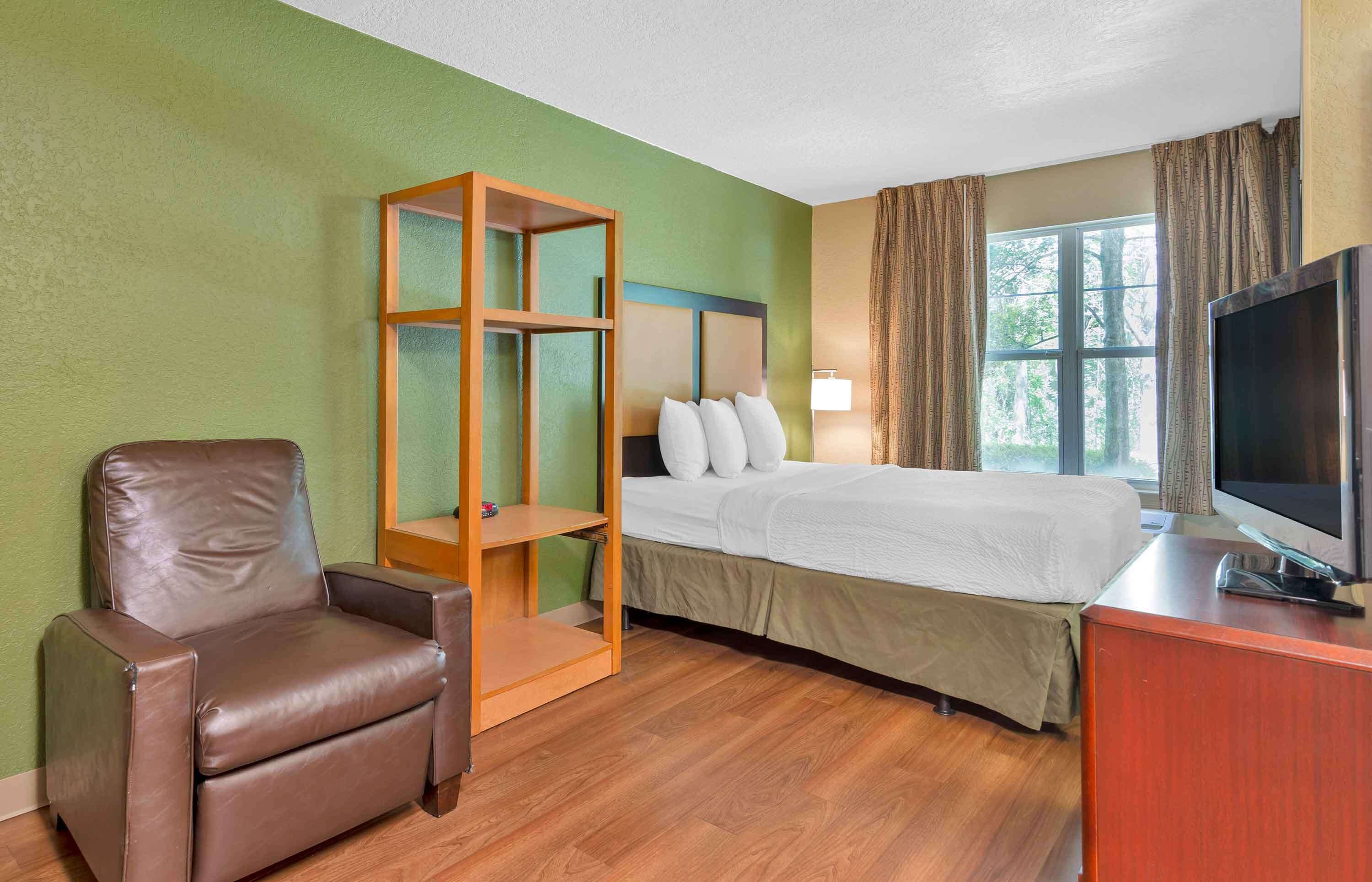 Una camera d'albergo con un letto, una televisione, una sedia e un mobiletto a mensole in legno.