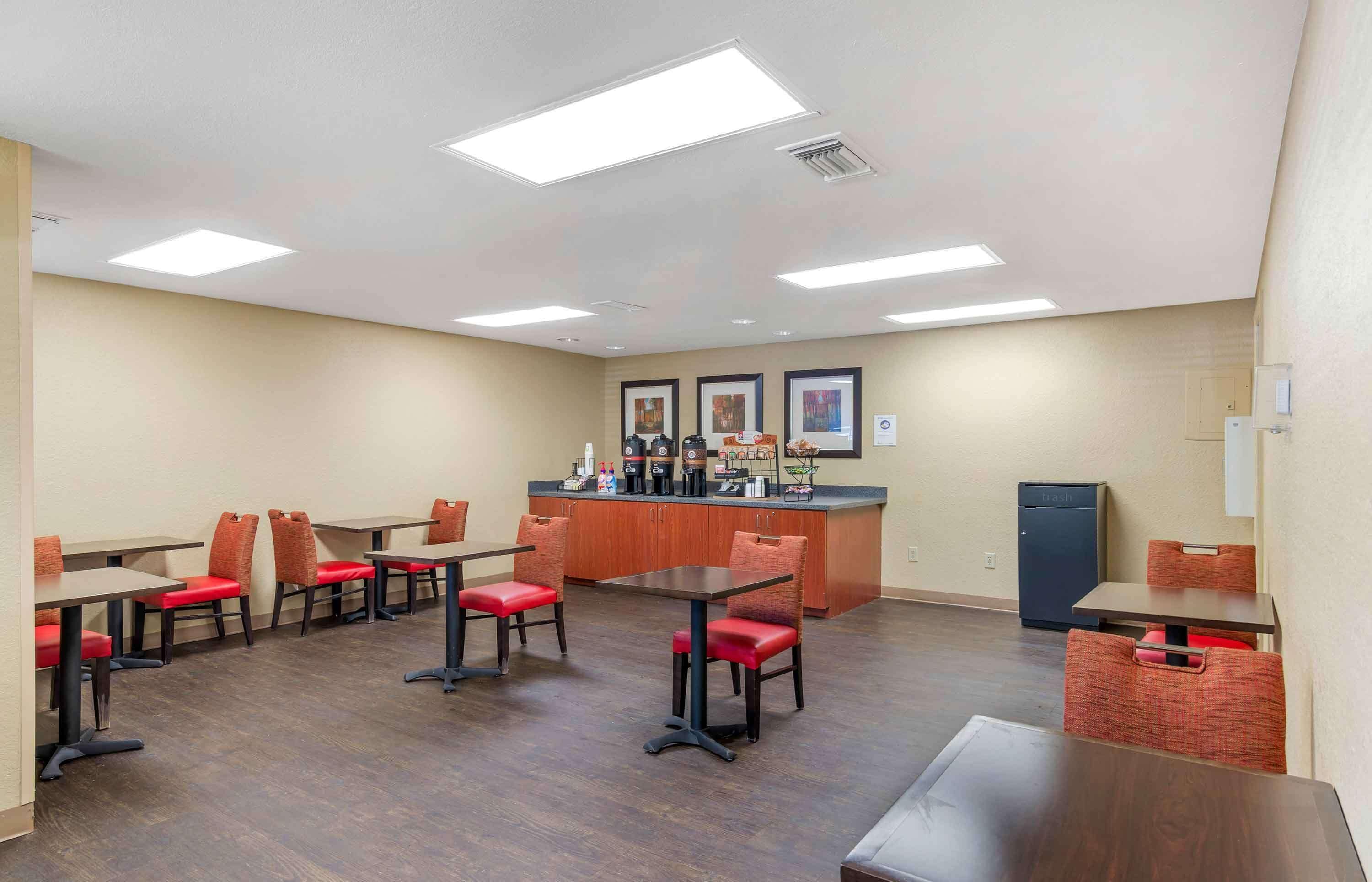A hotel dining area with multiple tables and chairs, a kitchen counter with appliances, and framed pictures on the wall.