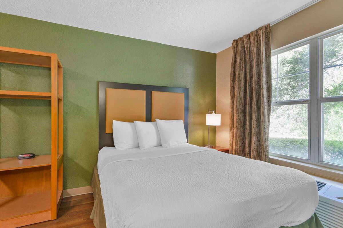 A hotel room with a bed, a wooden shelf, a window with curtains, and a nightstand with a lamp.