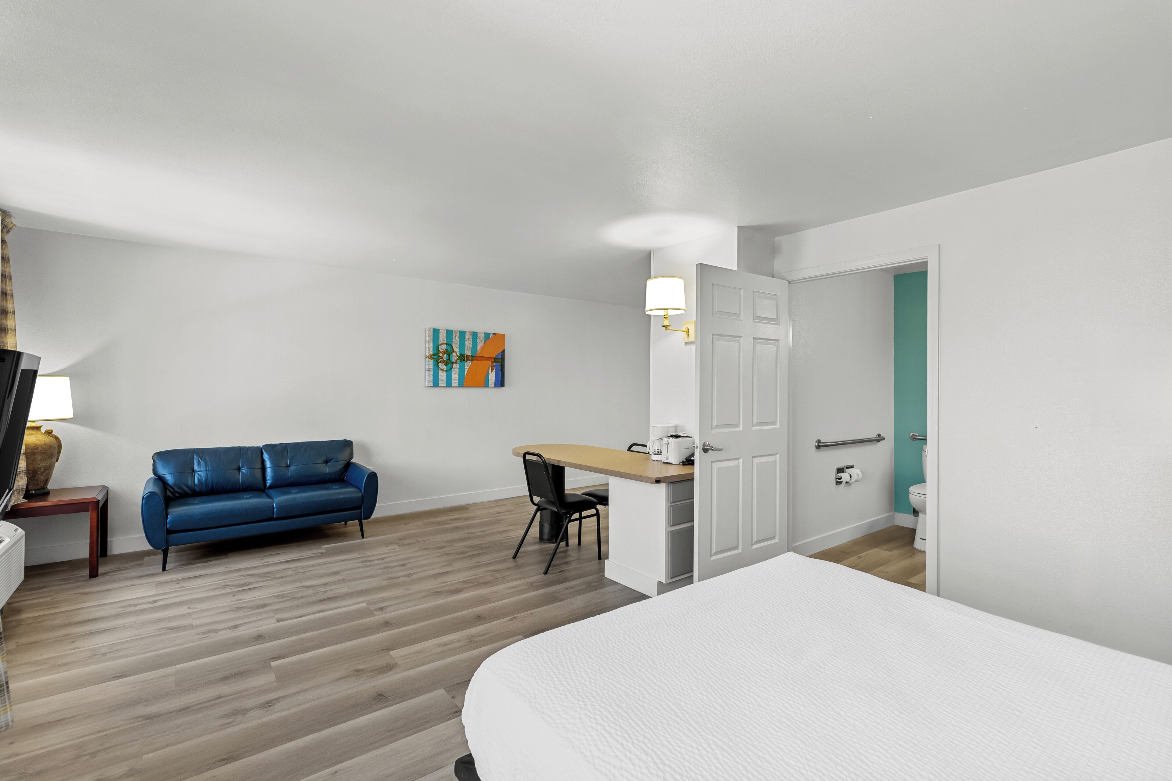Habitación de hotel con cama, sofá azul, escritorio con silla, mesita de noche con lámpara y puerta que da a un baño.