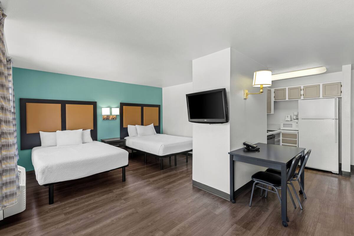Une chambre d’hôtel avec deux lits, une télévision, une table à manger avec des chaises, un coin cuisine et une salle de bain avec douche.