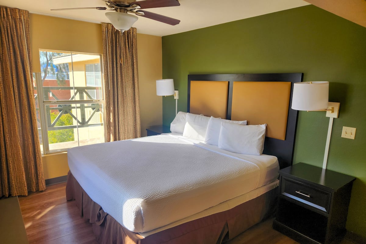 A neatly made bed with a headboard, two bedside lamps, a nightstand, and a window with curtains.