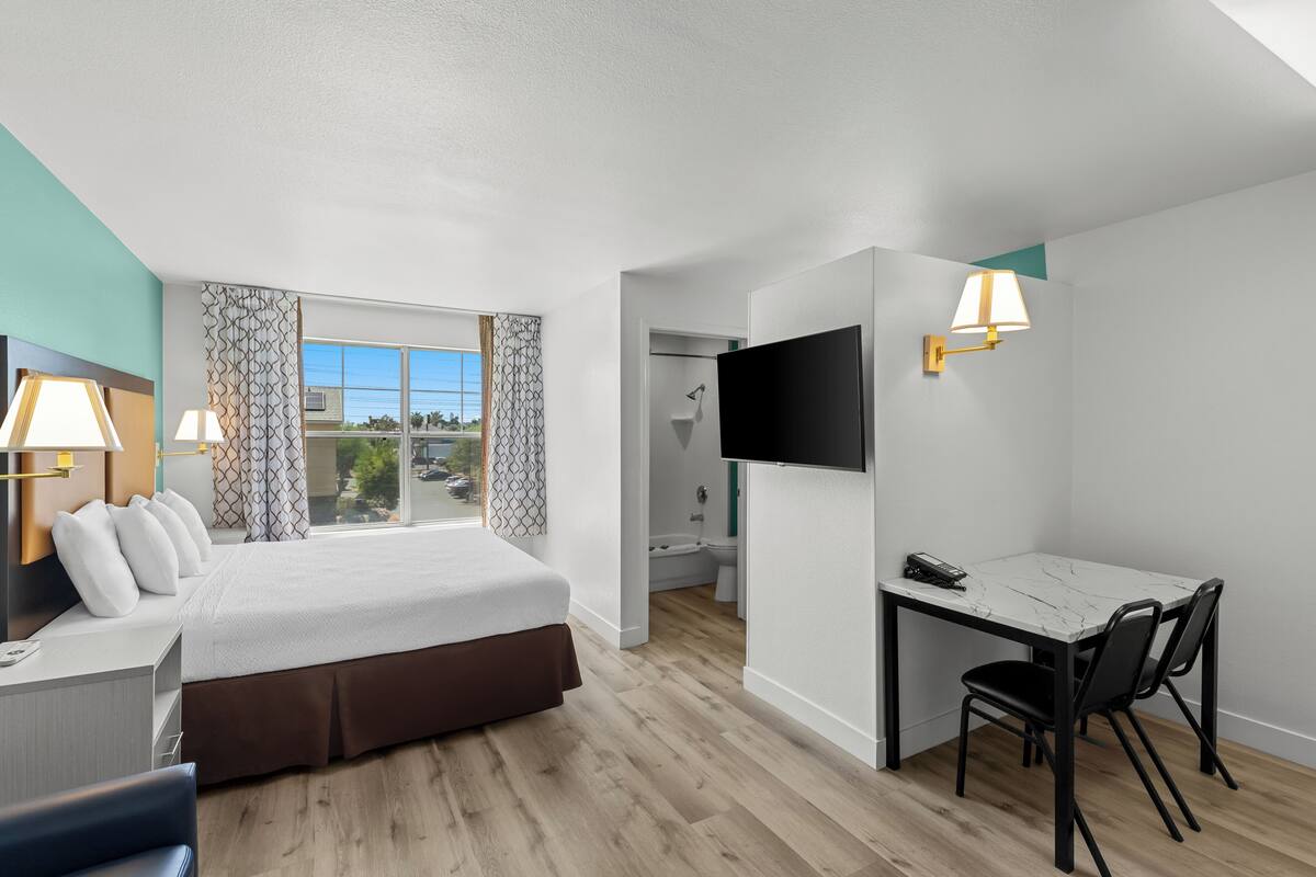 Une chambre d’hôtel avec un lit, un bureau, des chaises, une télévision et une salle de bain visible à travers une porte ouverte.