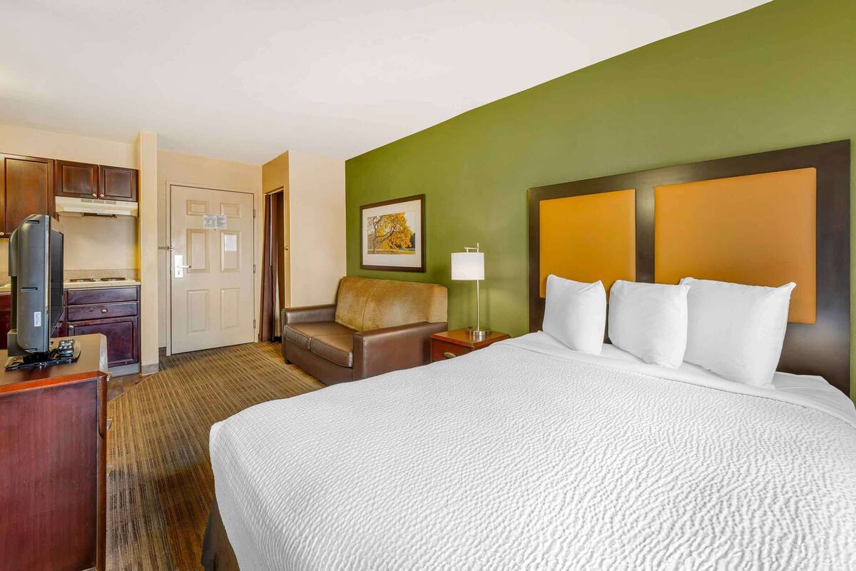 Ein Hotelzimmer mit einem großen Bett, einem Schreibtisch mit Computer, einem Sessel, einem Fernseher und einem Gemälde an der Wand.