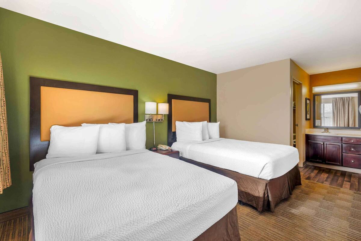 Ein Hotelzimmer mit zwei Betten, einem kleinen Schreibtisch mit Lampe, einem Spiegel und einem Fenster mit Vorhängen.