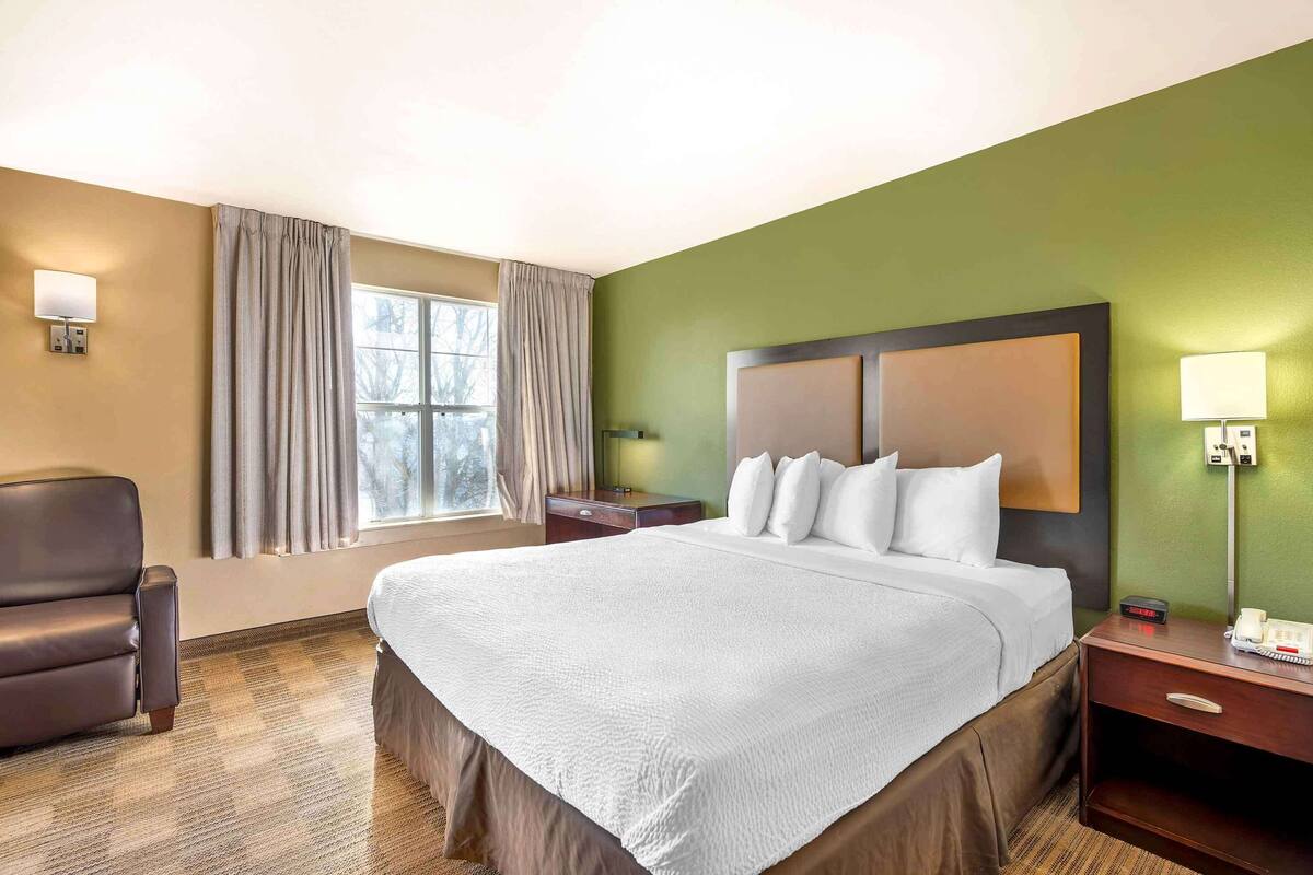 Ein Hotelzimmer mit einem großen Bett, einem Nachttisch mit Telefon, einer Lampe, einem Sessel und einem Fenster mit Vorhängen.