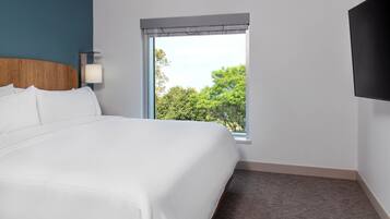 A hotel room with a large bed, a window with a view of trees, a wall-mounted TV, and a bedside lamp.