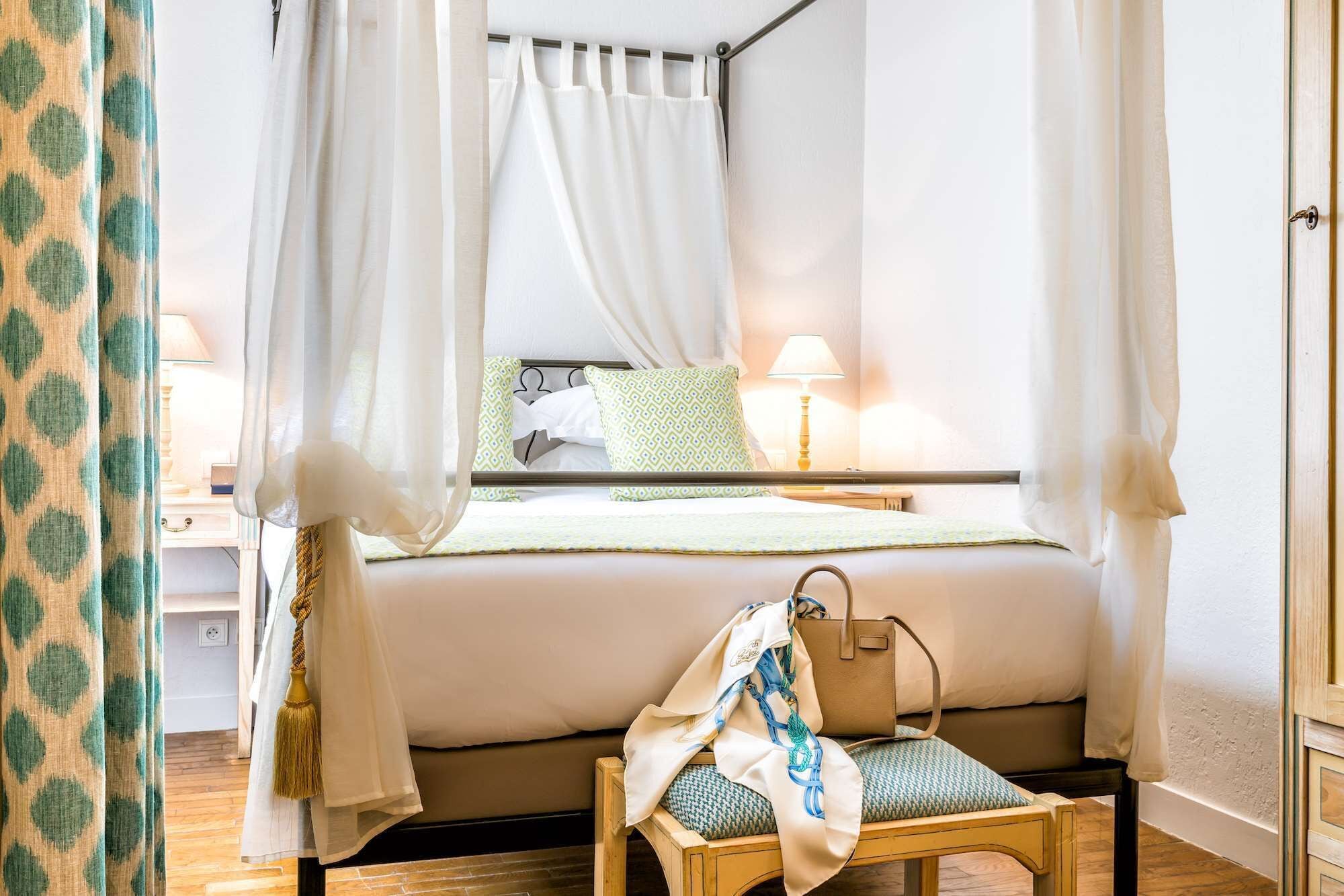 A bedroom with a four-poster bed, a bedside table with a lamp, and a chair with a bag.