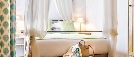 A bedroom with a four-poster bed, a bedside table with a lamp, and a chair with a bag.