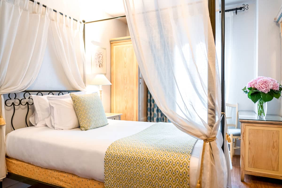 A neatly made bed with a patterned bedspread, a bedside table with a lamp, and a wooden nightstand with a vase of flowers.