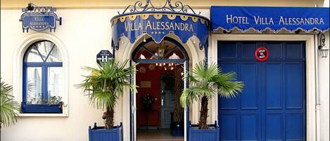 Exterior del Hotel Villa Alessandra, con fachada azul, un letrero sobre la entrada y plantas en macetas a la entrada.