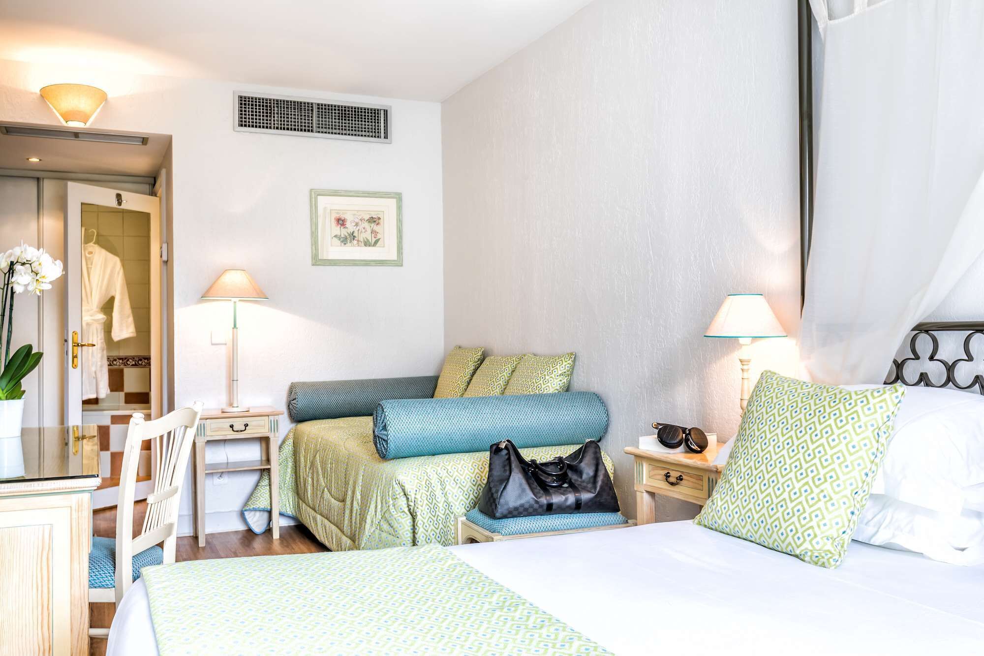 A hotel room with a bed, two bedside tables, a chair, a desk, a lamp, a picture frame, and a bathroom visible through an open door.