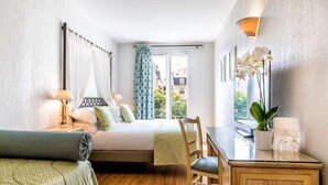 Habitación de hotel con cama, escritorio, silla, lámpara, planta en maceta y vistas al exterior.