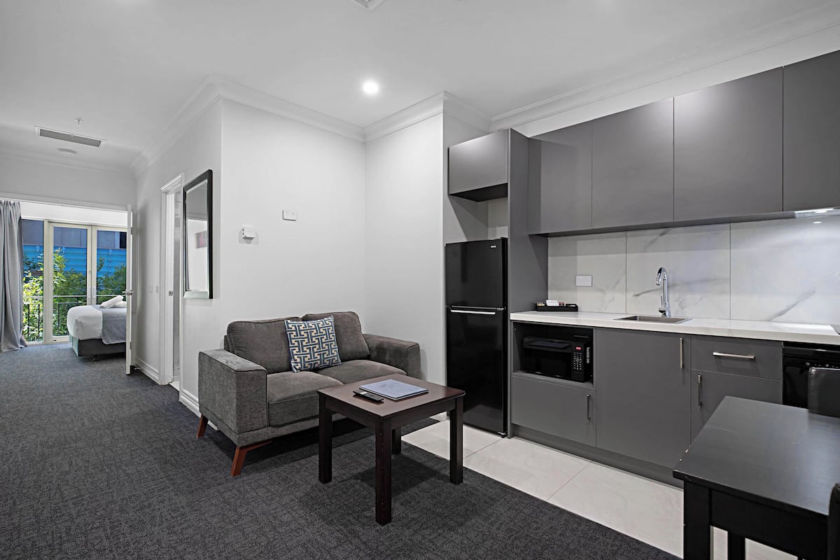 A modern apartment with a grey sofa, a small table, a kitchen with grey cabinets, and a balcony with a view.
