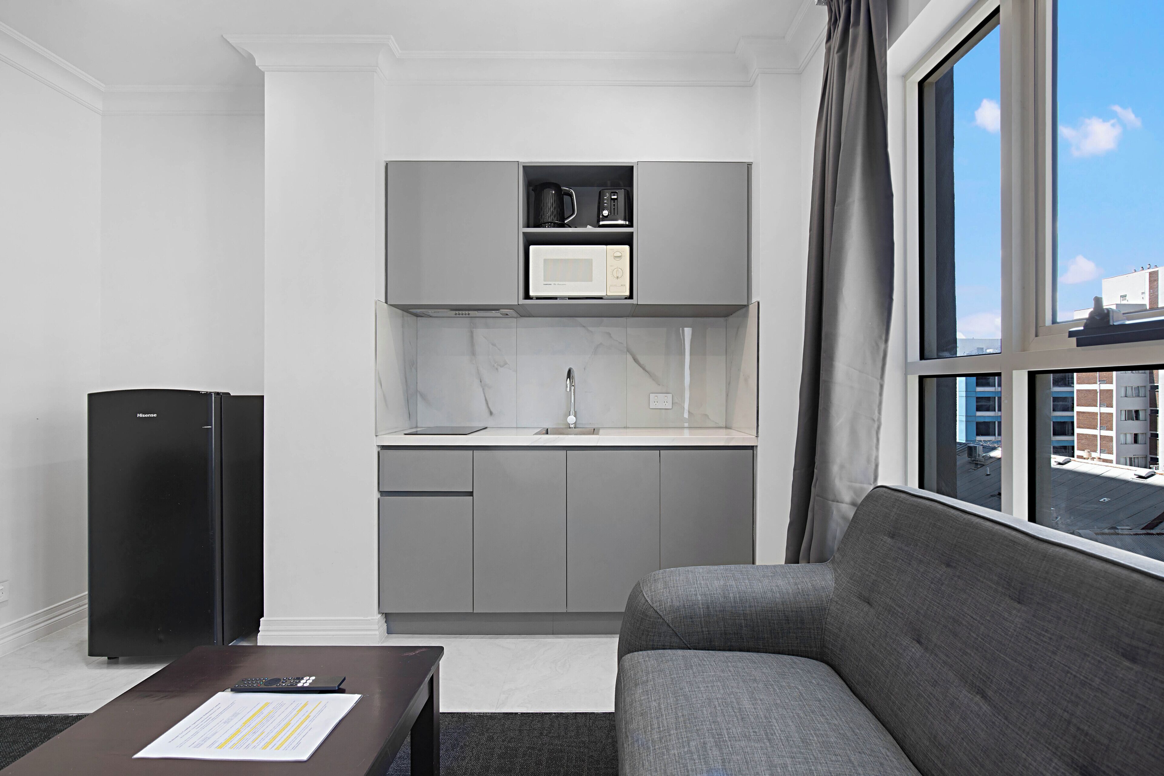 A compact kitchen with a microwave, coffee maker, and refrigerator, a gray sofa, and a window with a city view.