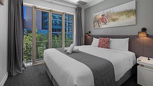 Un cuarto de hotel con una cama grande, vista de un edificio a través de una puerta corrediza de vidrio, un cuadro en la pared y una mesita de noche con una lámpara.
