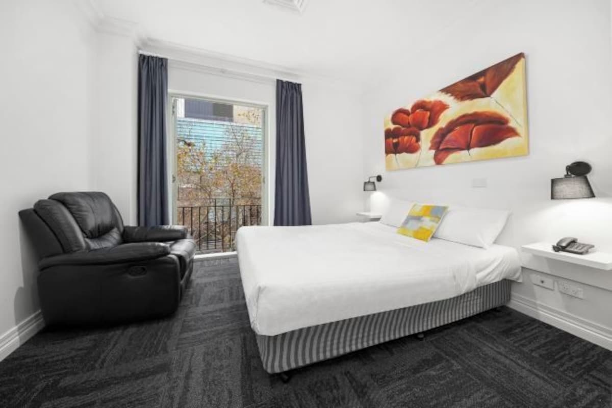 Habitación de hotel con cama, sofá de cuero negro, mesita de noche con lámpara y ventana con cortinas.