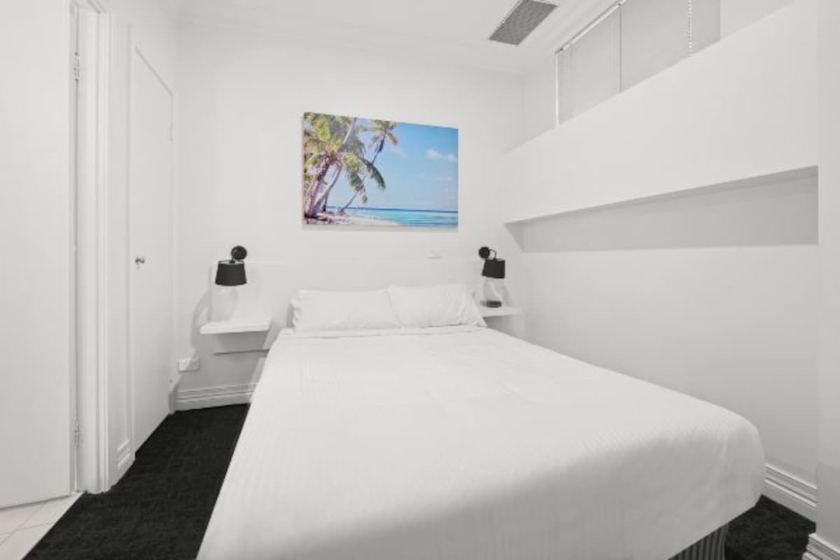 Un dormitorio con una cama, una mesita de noche, un cuadro de una playa y una bolsa negra sobre la mesita.
