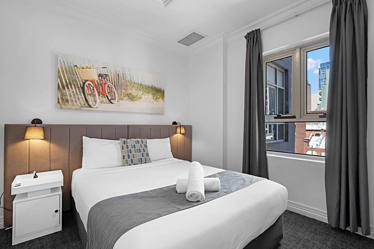 A hotel room with a bed, a nightstand, a lamp, a window with curtains, and a painting on the wall.