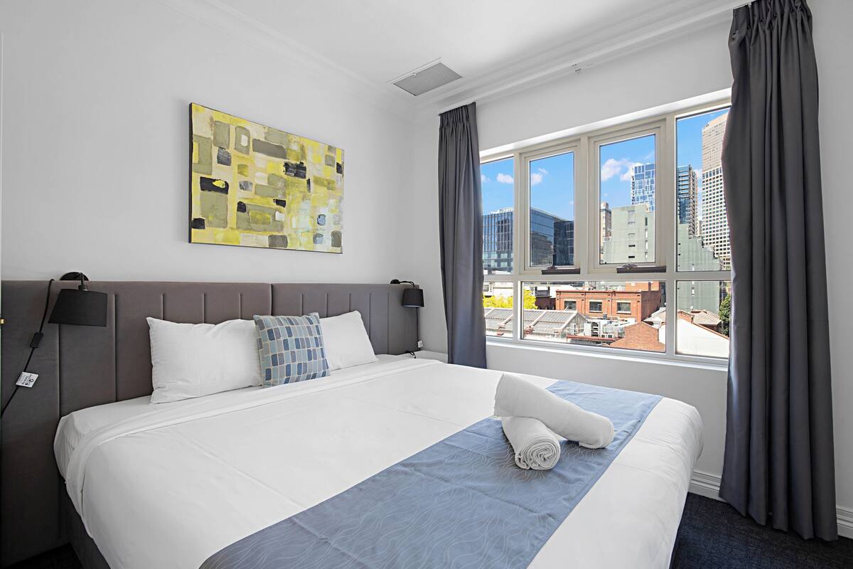 A hotel room with a large bed, a city view, a modern abstract painting, and a window with curtains.