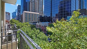 Un paisaje urbano con edificios altos modernos, un balcón con barandilla y abundante follaje verde.
