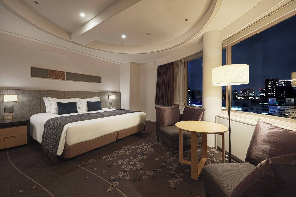 A hotel room with a large bed, a desk, a chair, and a view of the city at night.