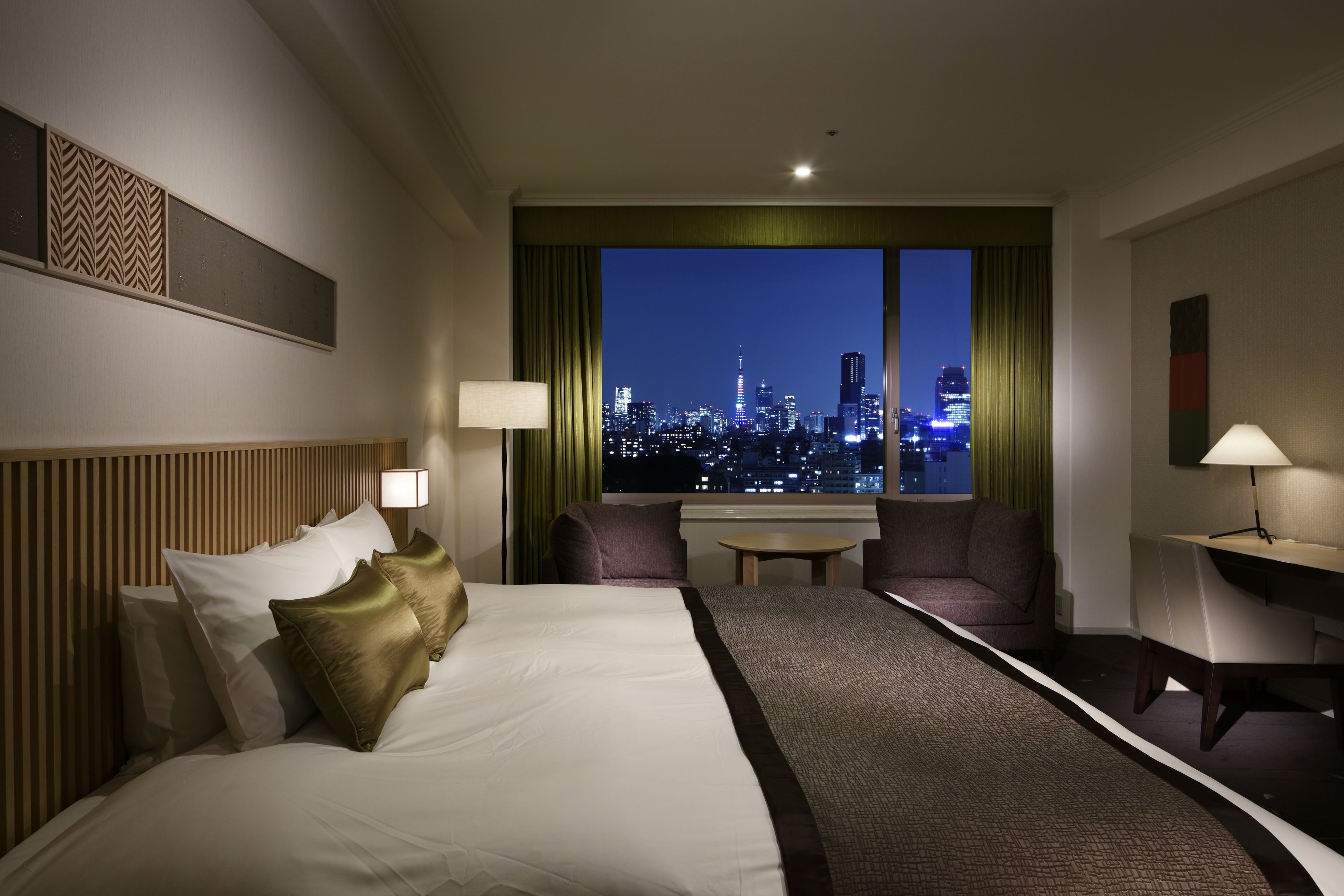 A hotel room with a large bed, two chairs, a small table, and a view of a cityscape at night.