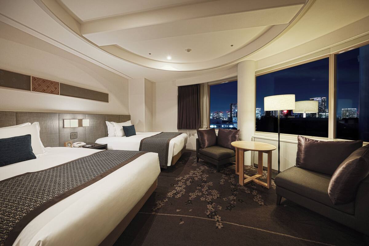 A hotel room with two beds, a sofa, a small table, and a view of the city at night.