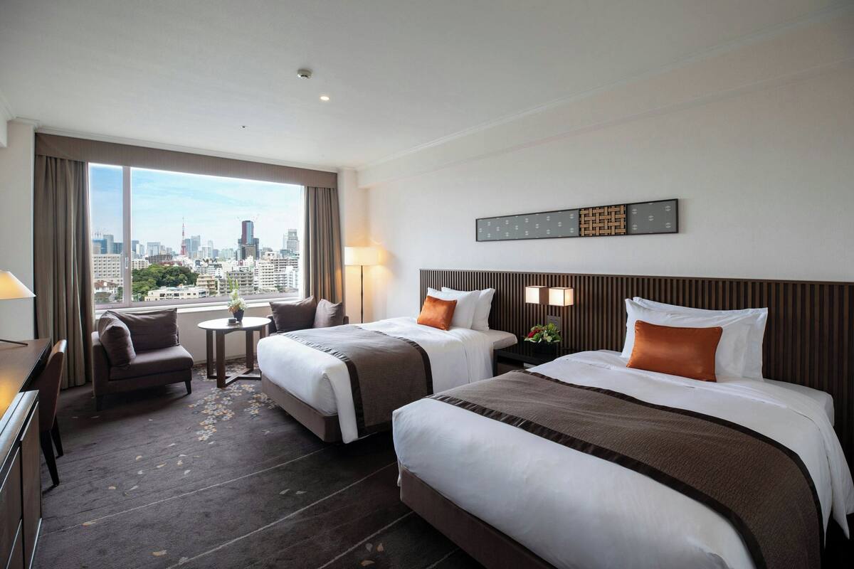 A hotel room with two beds, a desk, a chair, a small table with a lamp, and a city view.