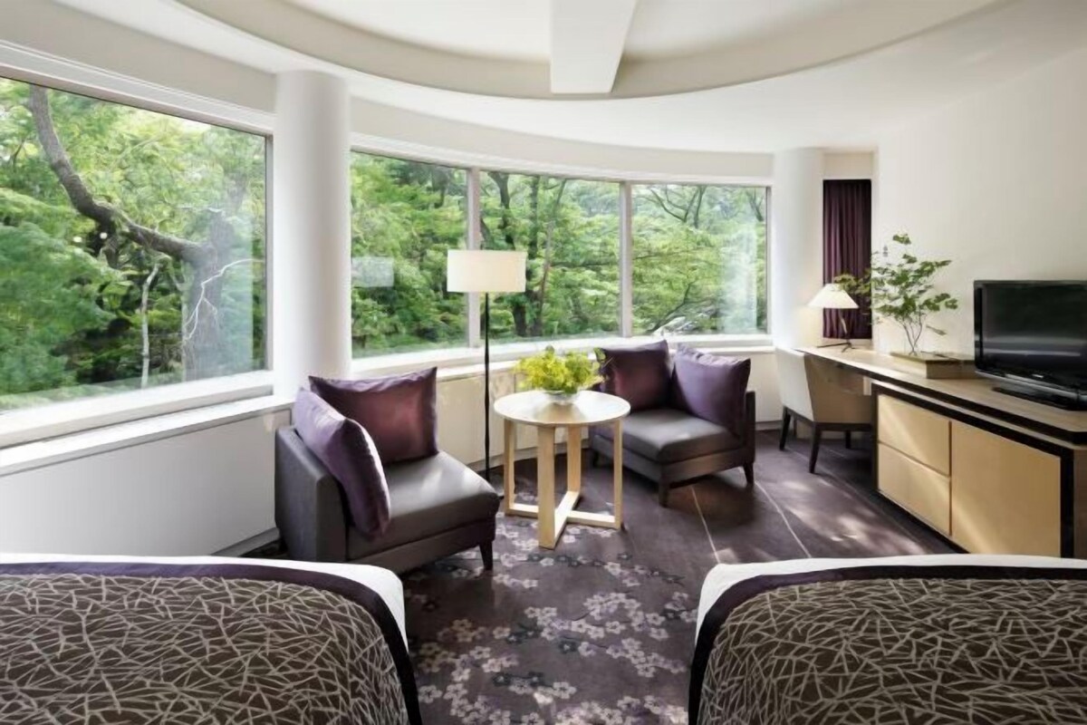 A modern hotel room with a curved ceiling, large windows with a forest view, a small round table with a lamp, and a TV on a cabinet.