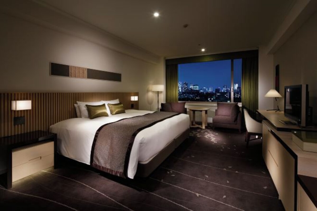 A hotel room with a large bed, a desk, a chair, and a view of a cityscape at night.