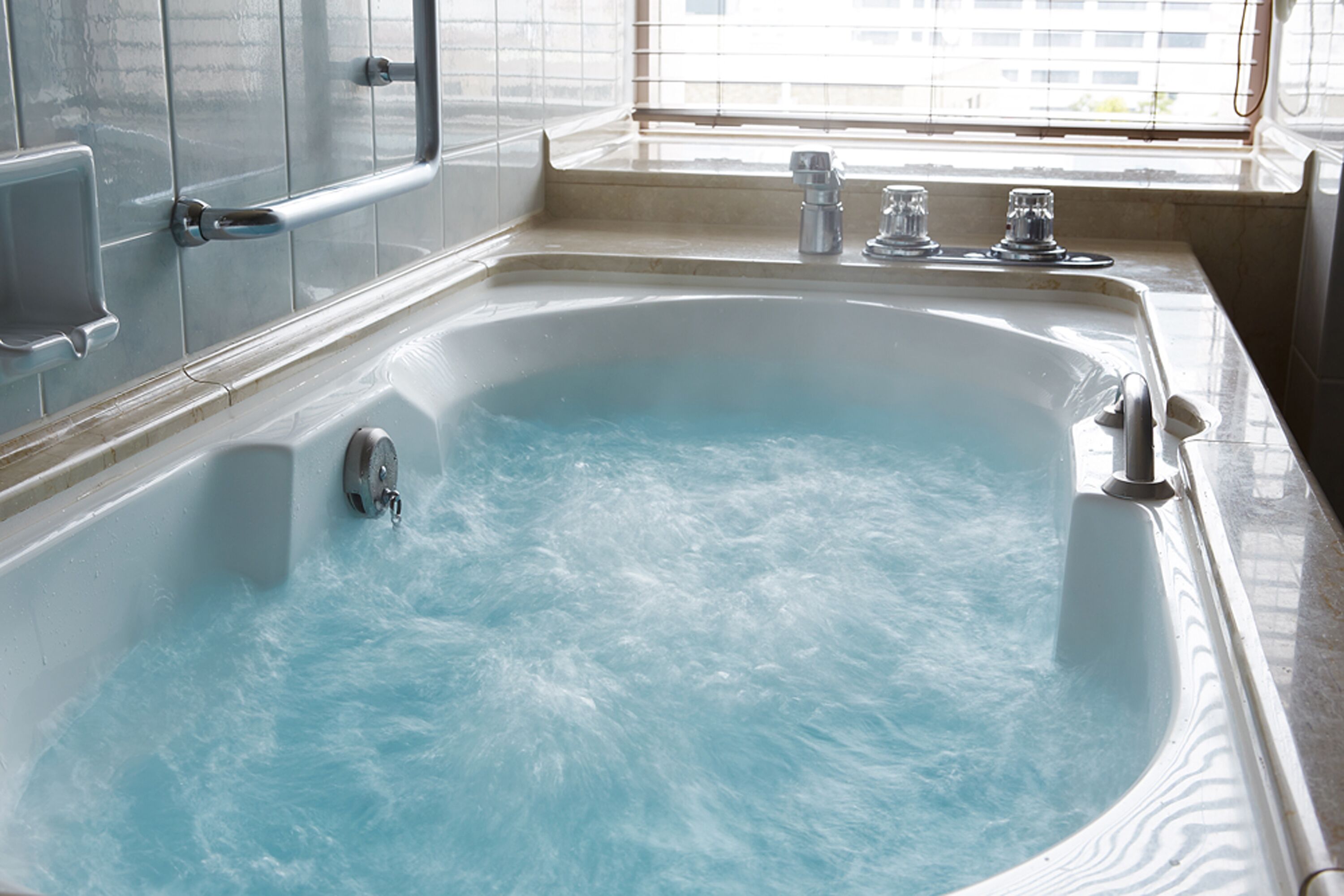 Une grande baignoire remplie d’eau, une fenêtre avec des stores et un mur carrelé.
