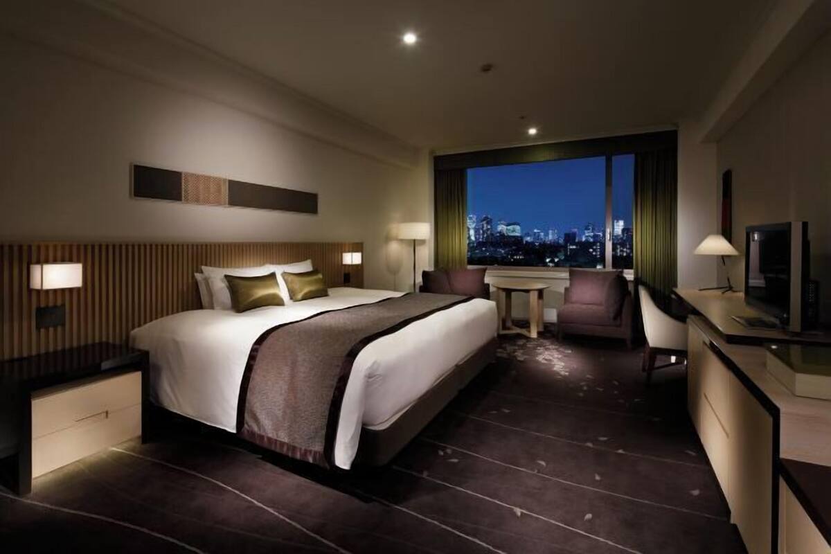 A hotel room with a large bed, a desk, a chair, and a view of a cityscape at night.