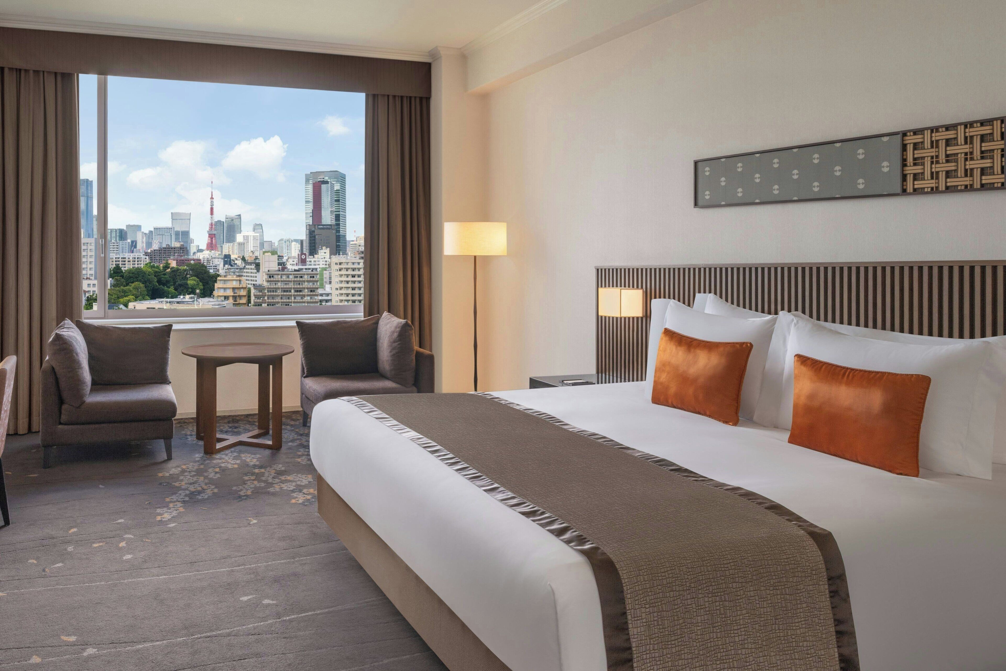 Ett hotellrum med en stor säng, två fåtöljer, ett litet bord och utsikt över stadens silhuett.