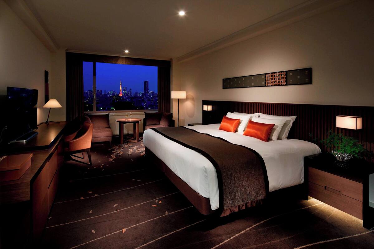 A hotel room with a large bed, a desk, a chair, a TV, and a view of a cityscape at dusk.